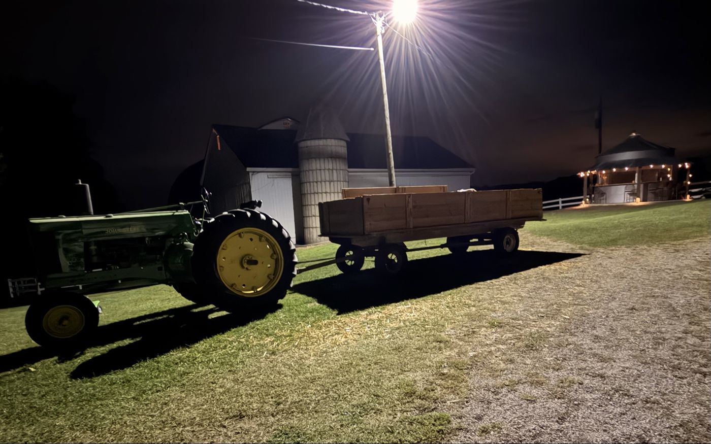 The lit grain bin and barnyard at night, with a green tractor and hay wagon